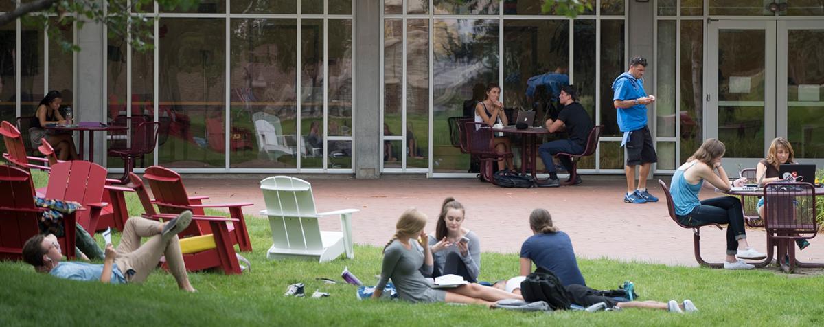 一群学生懒洋洋地躺在草地上，坐在玻璃墙外的桌子旁